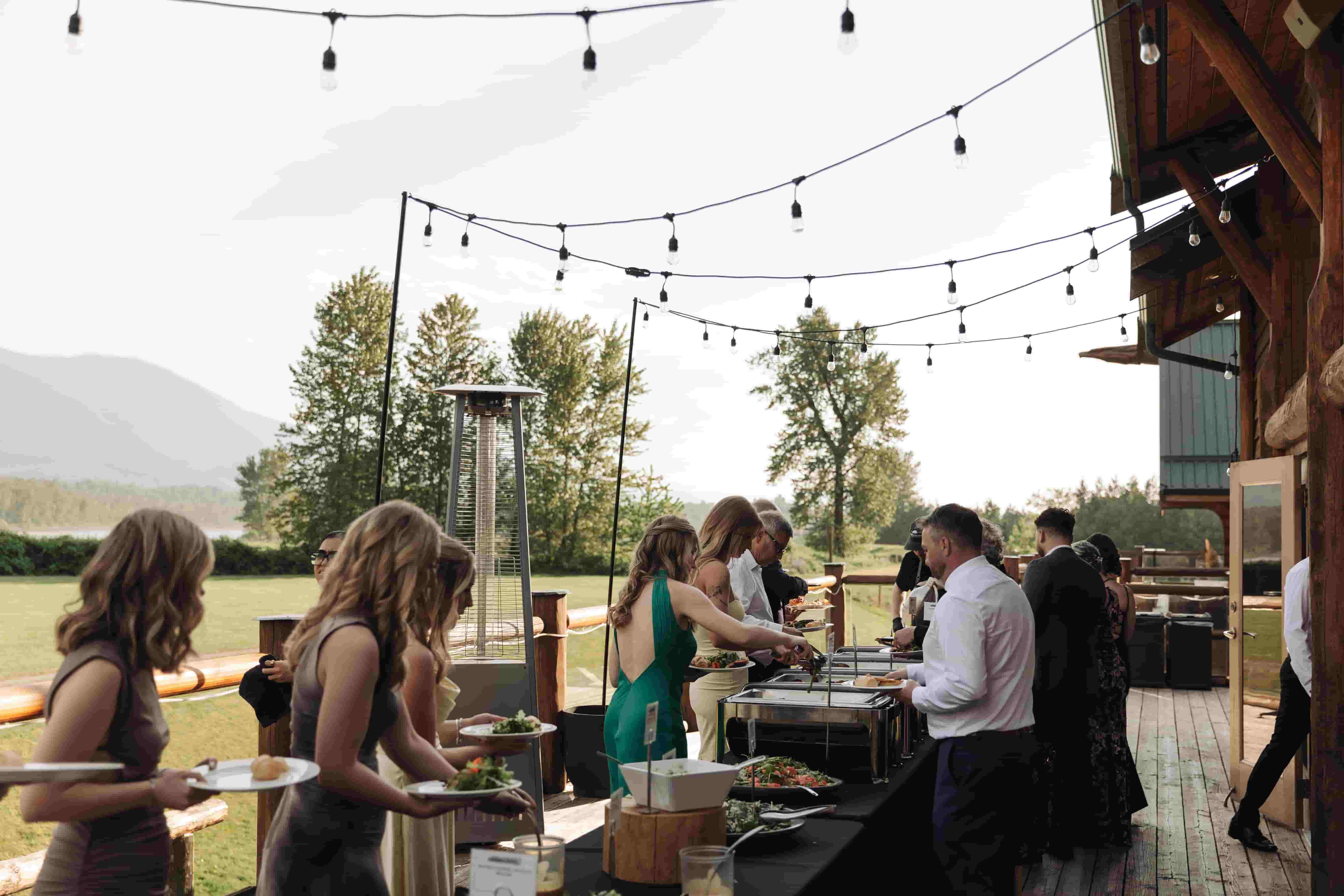 Wedding guests serving themselves at a buffet dinner on the Fraser River Lodge deck under string lights with mountain views