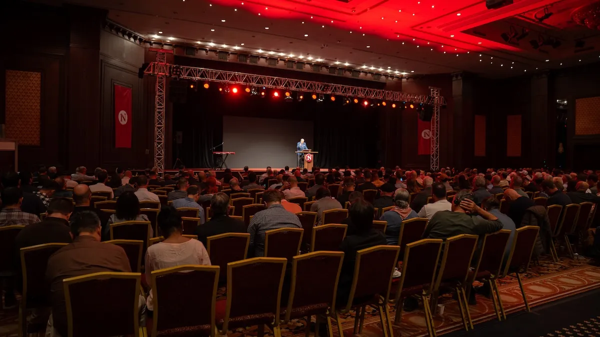 A large audience seated in rows facing a stage where a single speaker stands behind a podium under red stage lights in a conference hall.