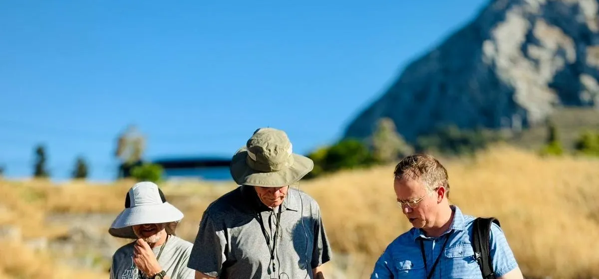 Three travelers in sun hats walking through the ruins of ancient Corinth, with Acrocorinth behind