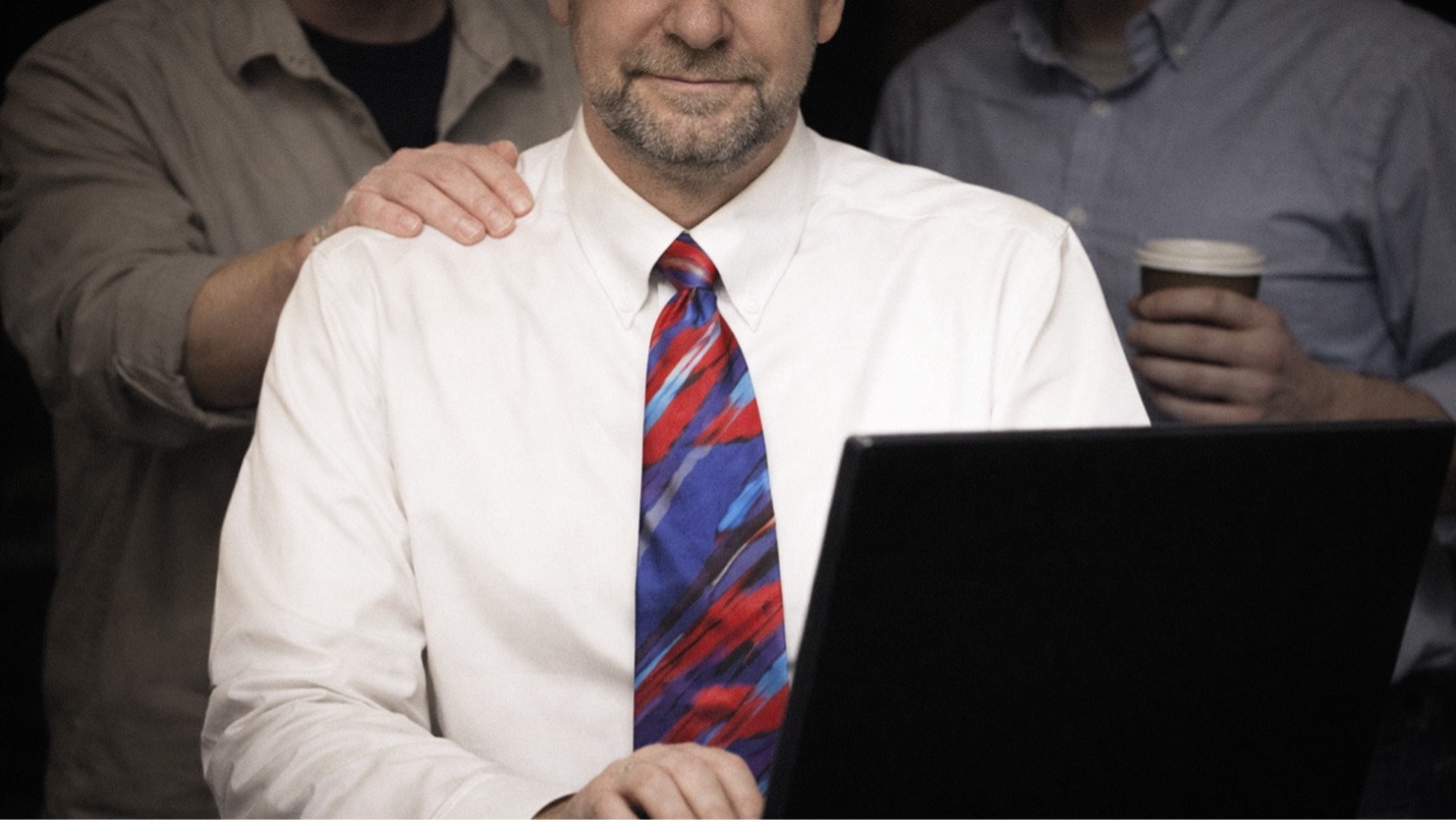 A Tech Lead sitting at a desk with a pensive smile, while programmers in the background try to get his attention.