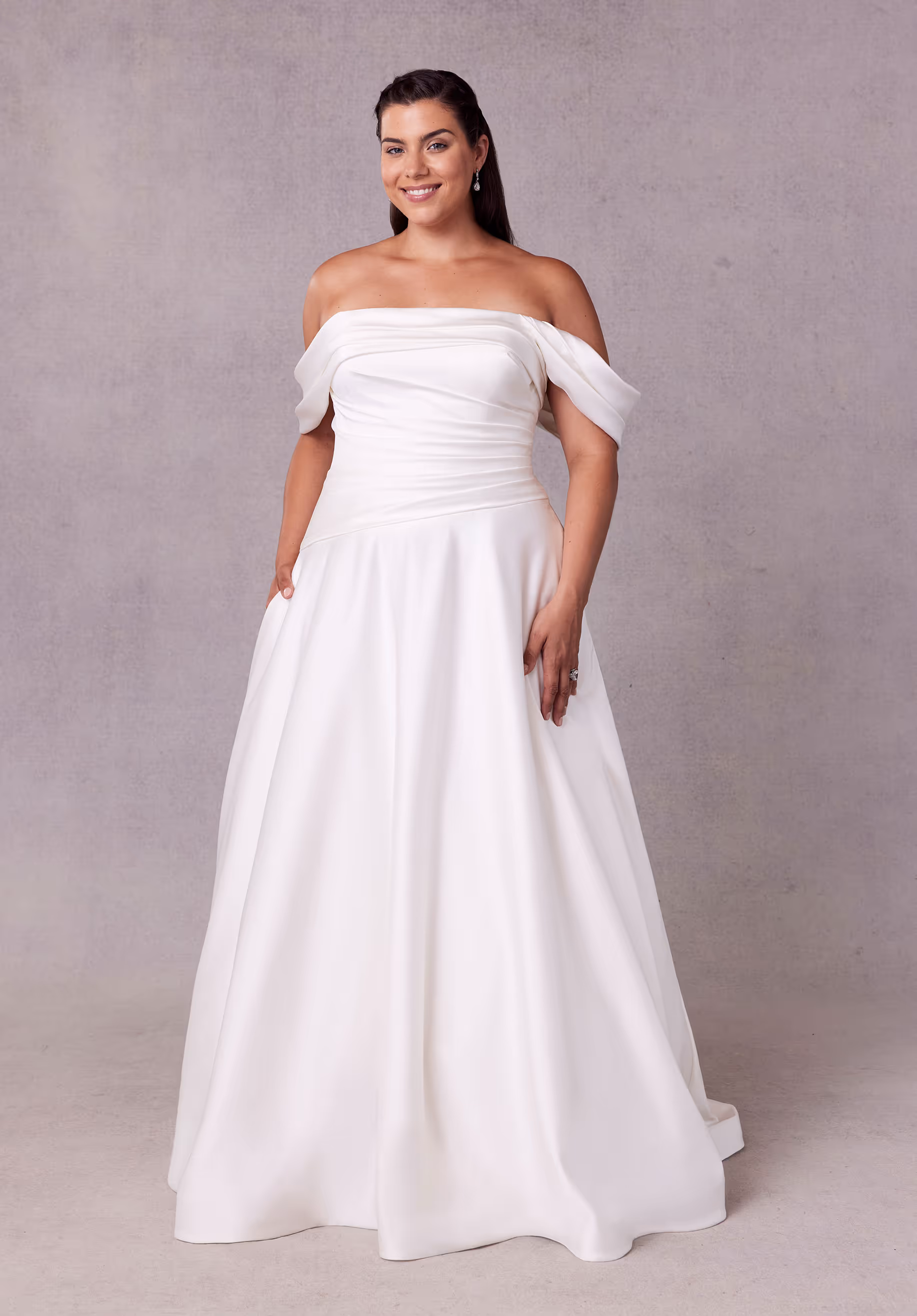 Understated satin wedding dress has an off-the-shoulder neckline with an asymmetrically draped bodice and waist on an A-line silhouette with a corset tie back.
