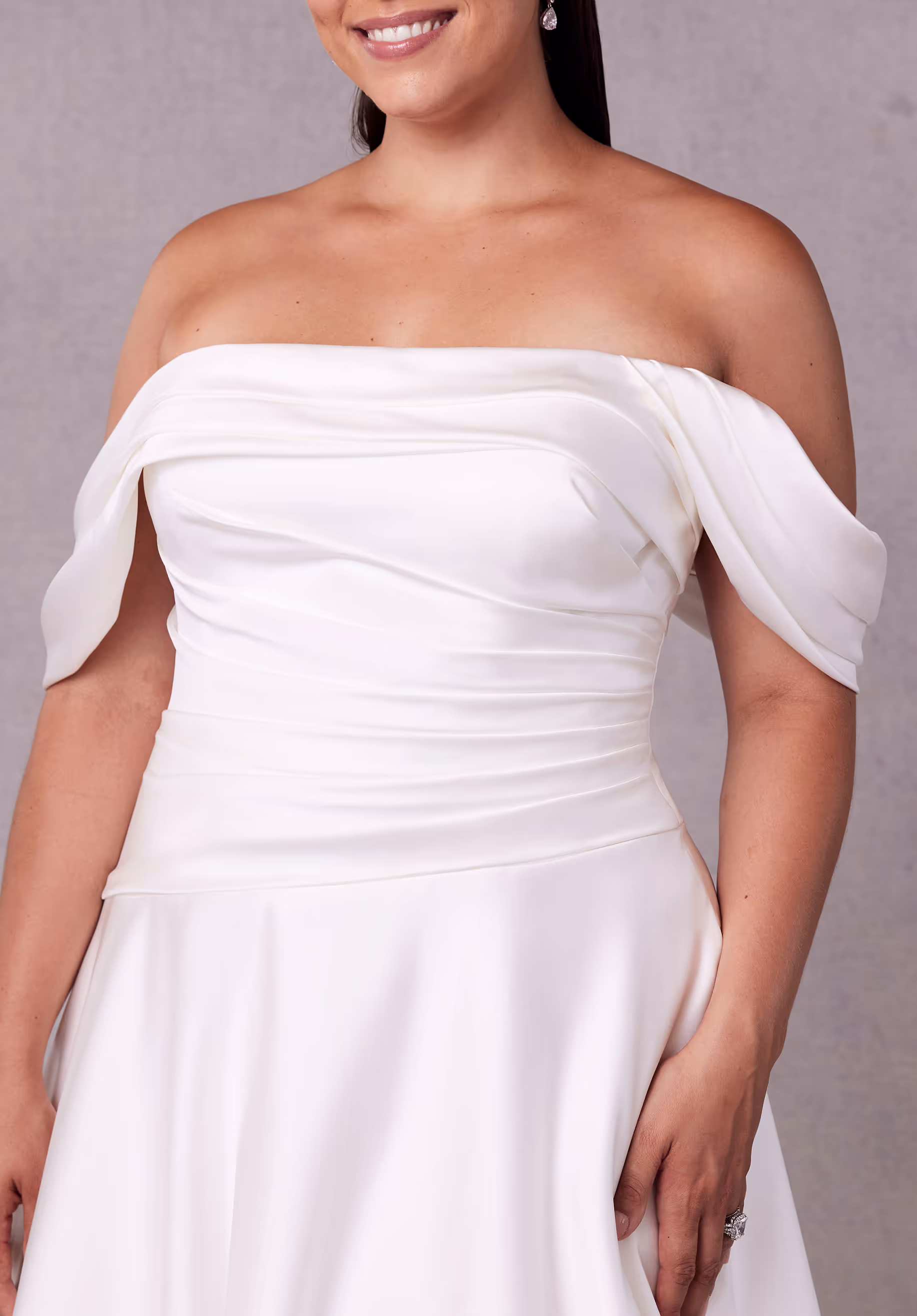 Understated satin wedding dress has an off-the-shoulder neckline with an asymmetrically draped bodice and waist on an A-line silhouette with a corset tie back.