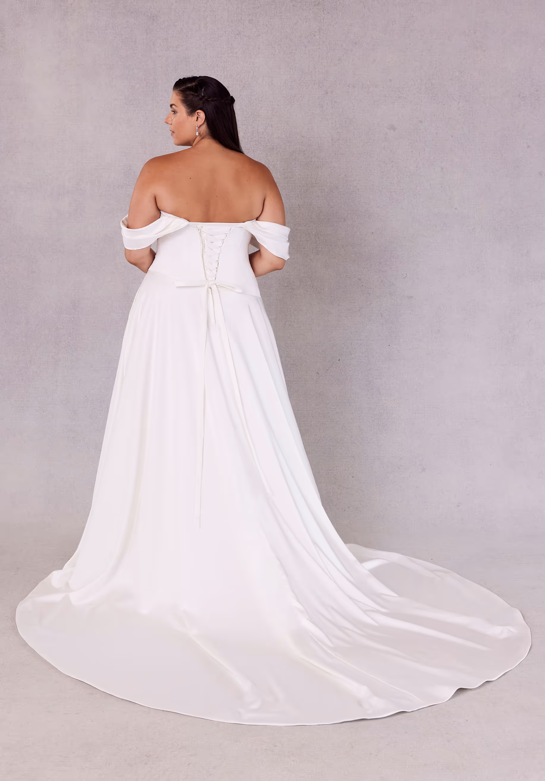 Understated satin wedding dress has an off-the-shoulder neckline with an asymmetrically draped bodice and waist on an A-line silhouette with a corset tie back.