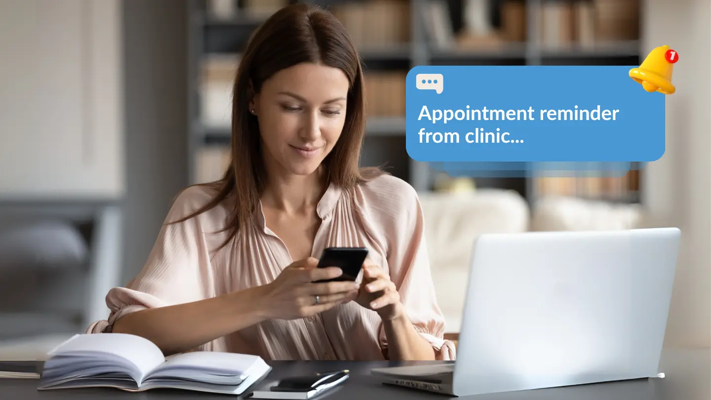 A woman sits at a desk with a laptop and open notebook, smiling as she looks at her smartphone. A digital notification graphic above her reads, “Appointment reminder from clinic…” with a bell icon showing one new alert. The setting is a cosy home or office environment with bookshelves in the background.