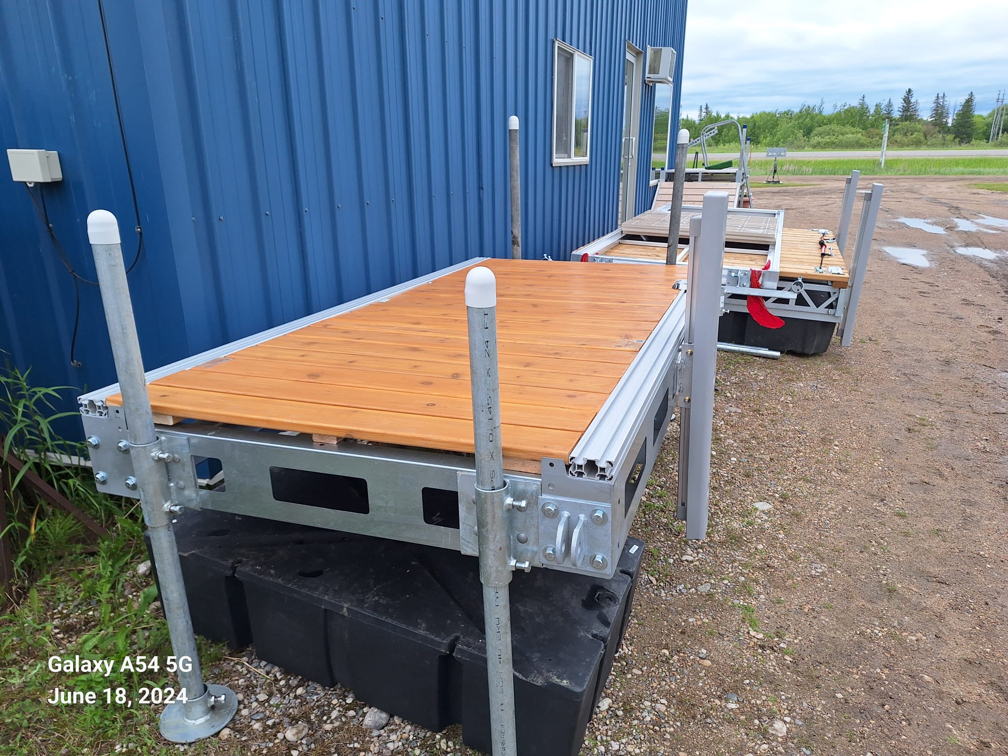 Two modular floating dock sections with wooden tops and metal frames next to a blue building on a gravel surface.