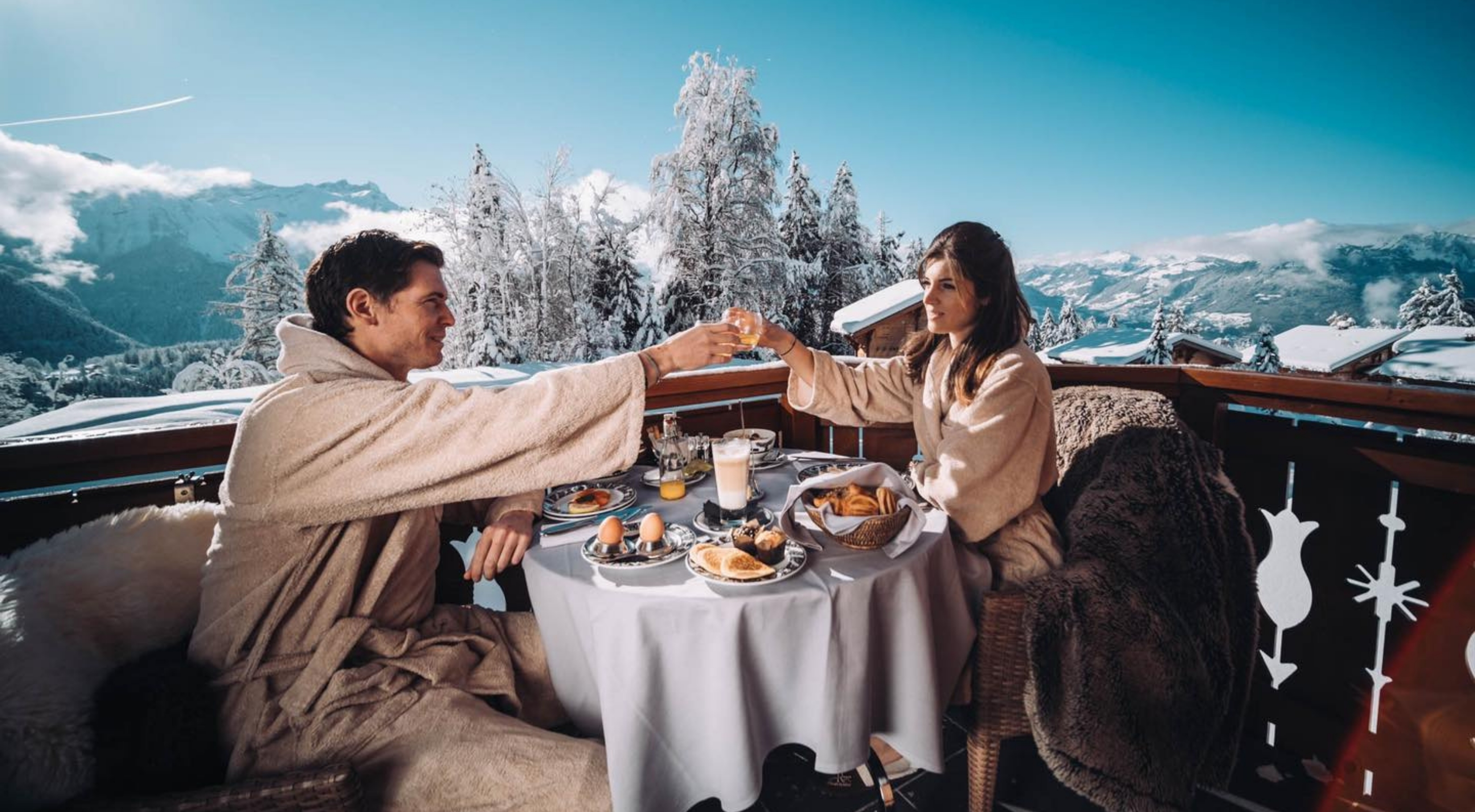 A couple toasts breakfast on the balcony of their room at Chalet Royalp in a snowy landscape.