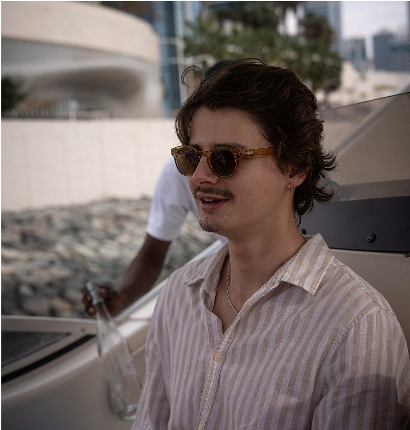 Un jeune homme portant des lunettes de soleil et une chemise rayée beige est assis sur un bateau, profitant d’une sortie en mer, avec des bâtiments modernes et des palmiers visibles en arrière-plan, créant une atmosphère estivale et détendue.
