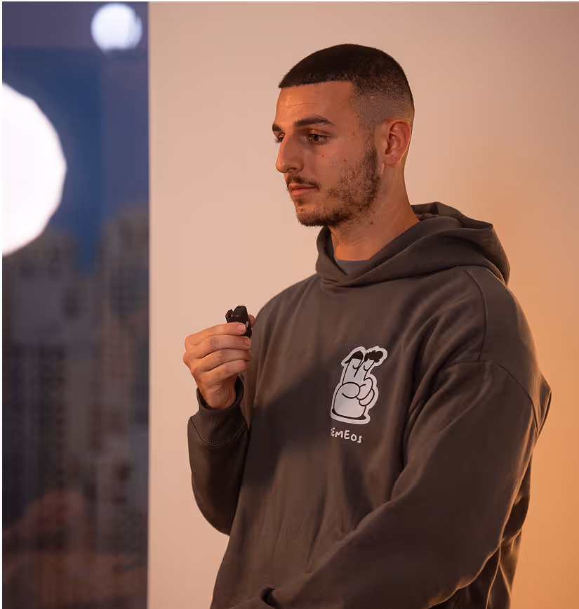 Un jeune homme en sweat à capuche gris avec un logo « GEMEOS » tient un micro pendant une présentation dans un appartement lumineux, avec en arrière-plan des immeubles de ville visibles à travers la fenêtre, créant une atmosphère professionnelle et détendue.
