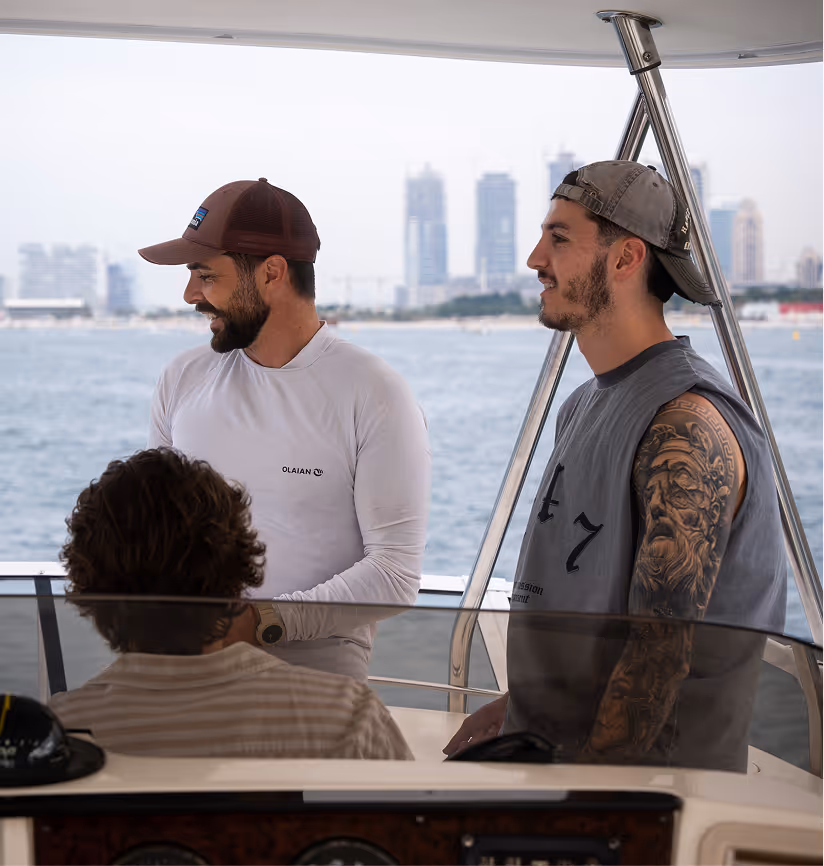 Trois hommes discutent à bord d’un bateau avec des immeubles modernes en arrière-plan, l’un portant un t-shirt à manches longues blanc, l’autre un débardeur gris laissant apparaître un grand tatouage sur son bras, et le troisième assis de dos devant le tableau de bord, sur fond de mer calme.
