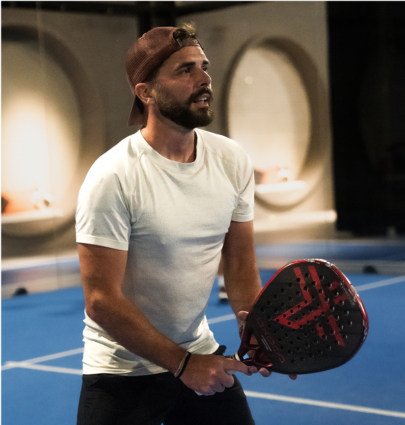 Un homme portant une casquette à l’envers et un t-shirt blanc se prépare à jouer au padel sur un court intérieur bleu, tenant sa raquette noire et rouge avec concentration avant de recevoir la balle.
