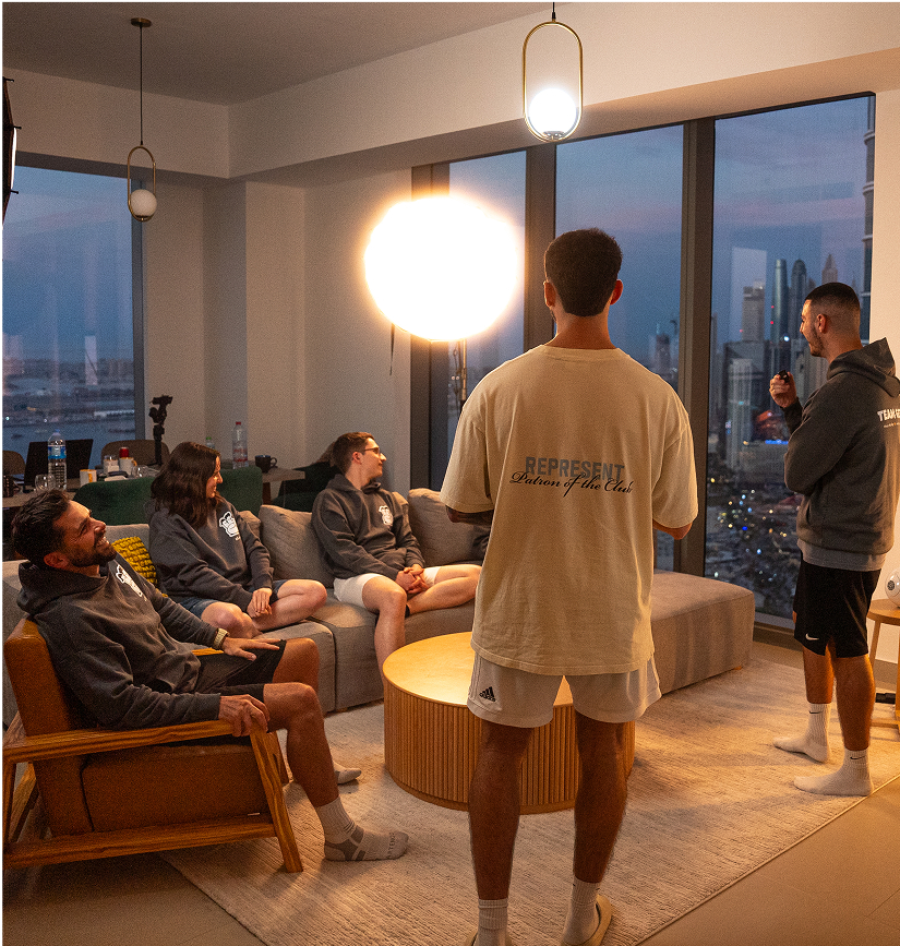 Cinq jeunes adultes discutent de manière détendue dans un salon moderne avec vue sur une ville illuminée, assis autour d’une table ronde en bois, certains en sweat à capuche gris, d’autres en t-shirt clair, sous un éclairage doux, créant une ambiance de réunion conviviale en soirée.
