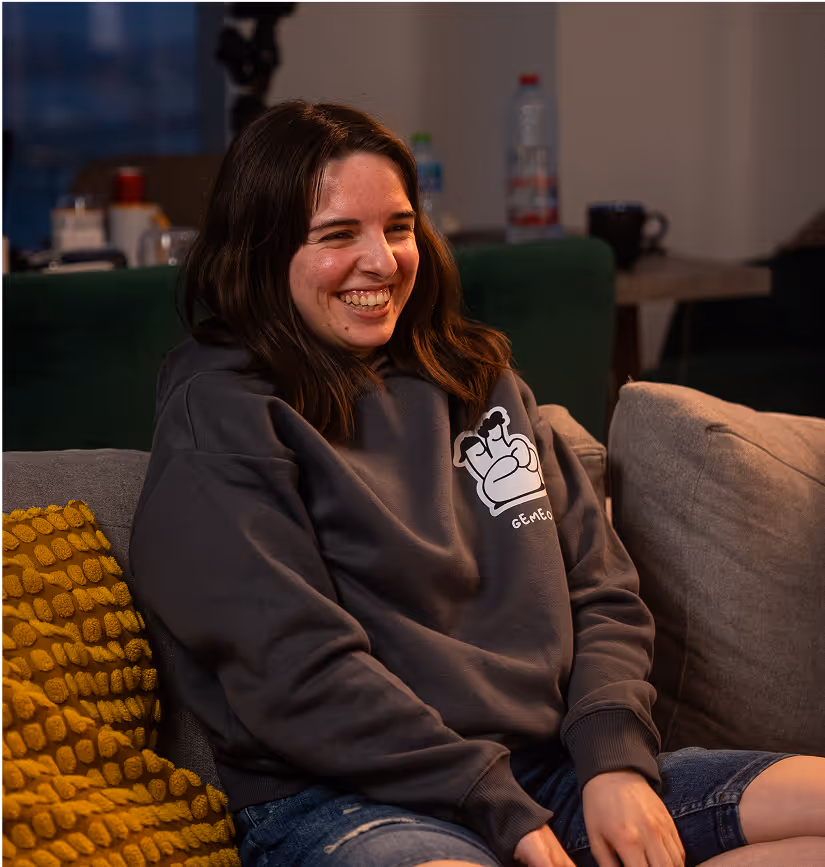 Une jeune femme assise sur un canapé, souriante, portant un sweat gris avec le logo « GEMEOS », profite d’un moment convivial dans un salon cosy, avec un coussin jaune texturé à ses côtés et un fond légèrement flou d’appartement en soirée.
