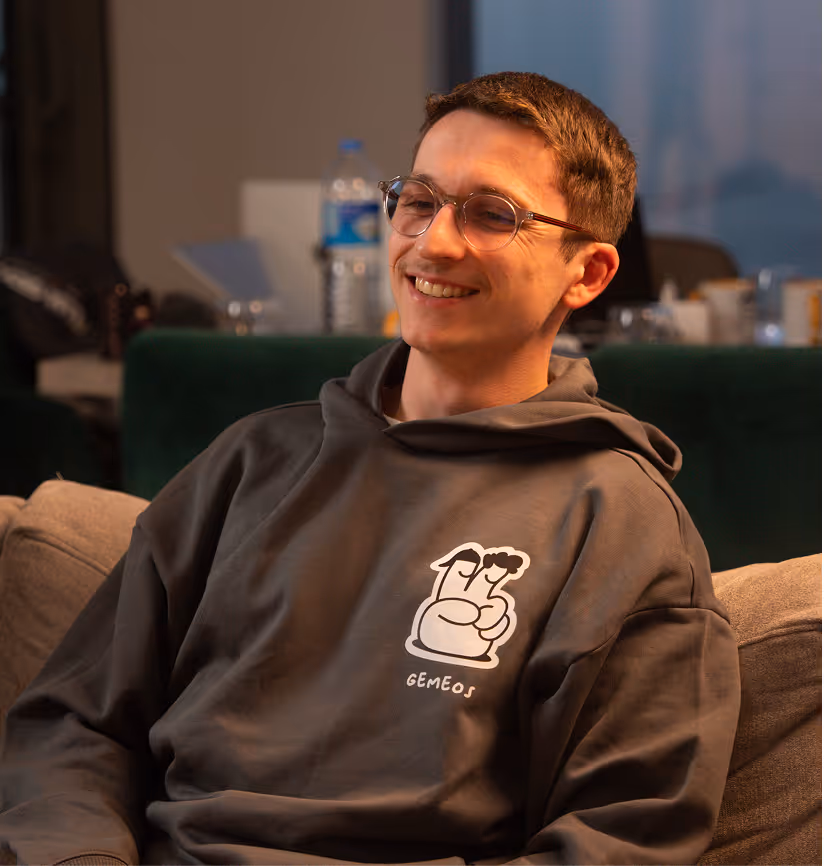Un jeune homme souriant, portant des lunettes et un sweat à capuche gris avec un dessin et le mot “GEMEOS”, est assis confortablement sur un canapé dans un intérieur lumineux, créant une atmosphère détendue et conviviale.
