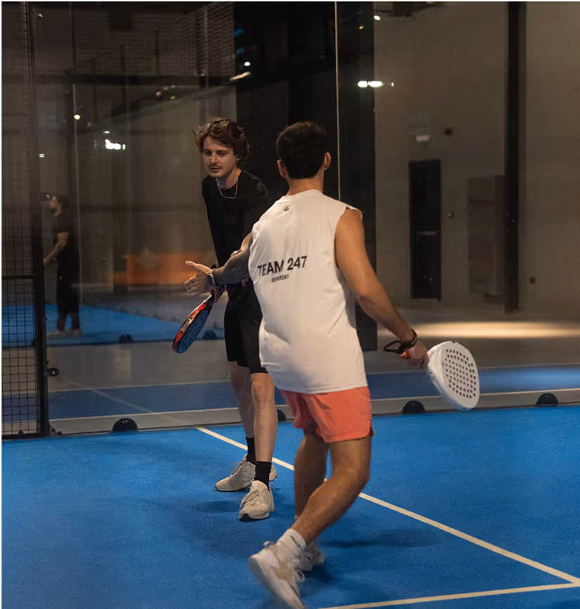 Deux joueurs de padel sur un court intérieur bleu échangent un geste de main après un point, l’un portant un t-shirt noir et l’autre un débardeur blanc marqué « TEAM 247 » avec un short rose, chacun tenant sa raquette, dans une atmosphère sportive et détendue.
