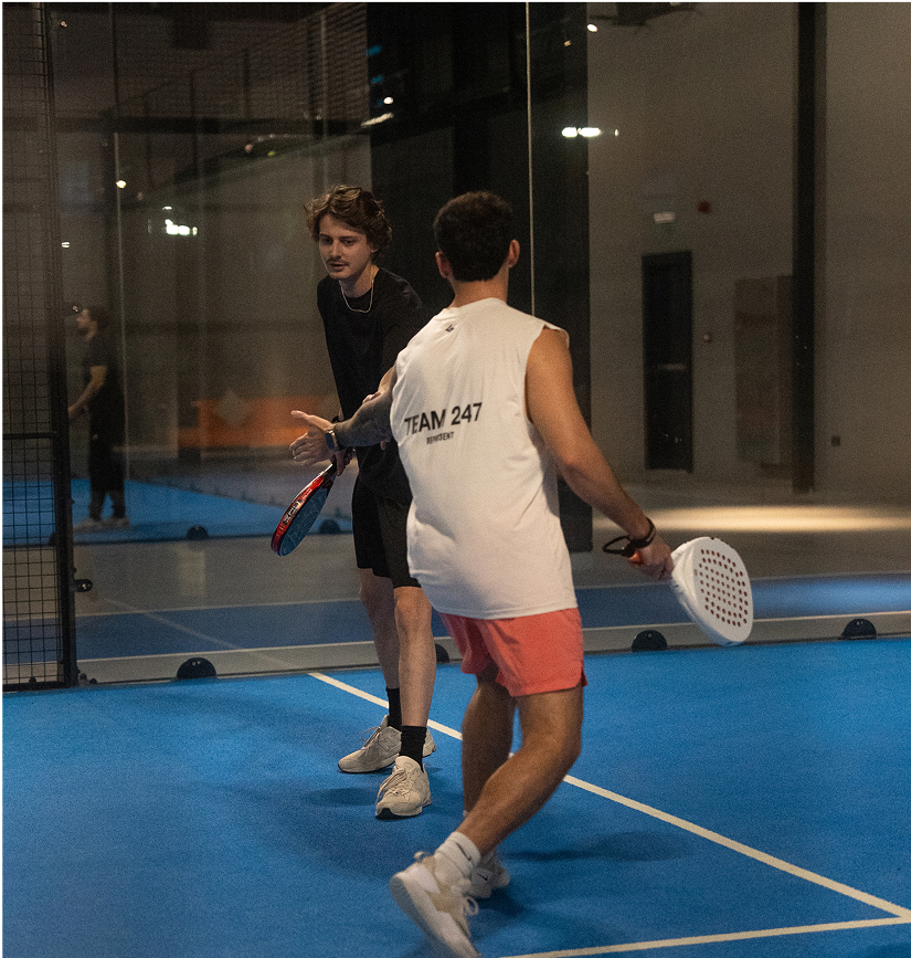 Deux joueurs de padel sur un court intérieur bleu échangent un geste de main après un point, l’un portant un t-shirt noir et l’autre un débardeur blanc marqué « TEAM 247 » avec un short rose, chacun tenant sa raquette, dans une atmosphère sportive et détendue.
