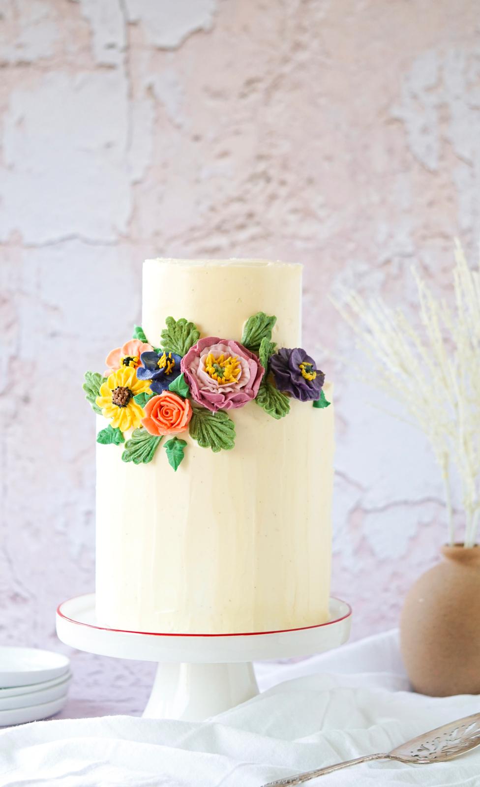 Vanilla cake with Swiss meringue buttercream topped with colorful fresh flowers on a white cake stand.