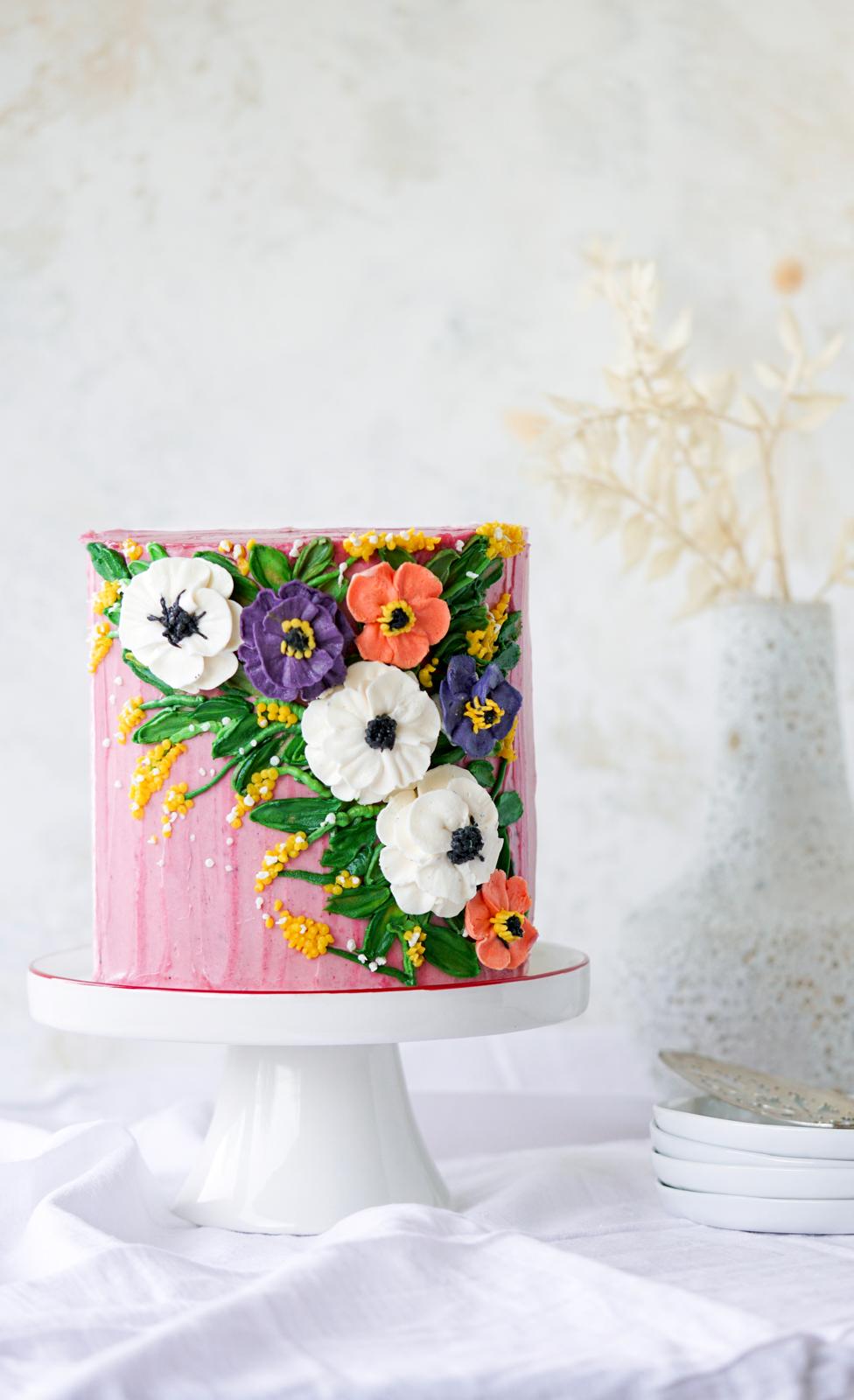 Pink buttercream cake with white, purple, orange, and yellow wildflower decorations on a white cake stand.