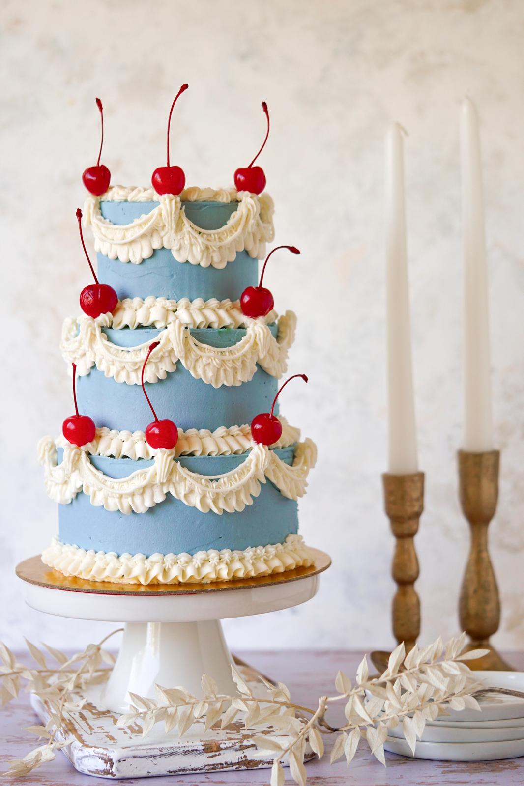 Three-tier blue cake decorated with ivory Lambeth-style piping and red cherries on top of each tier.