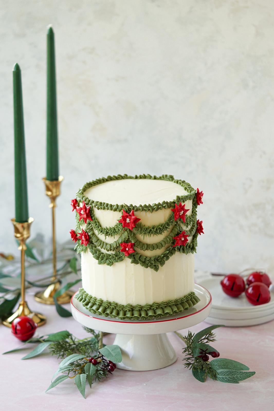 White buttercream cake decorated with green garlands, red poinsettias, and piped borders on a white cake stand.