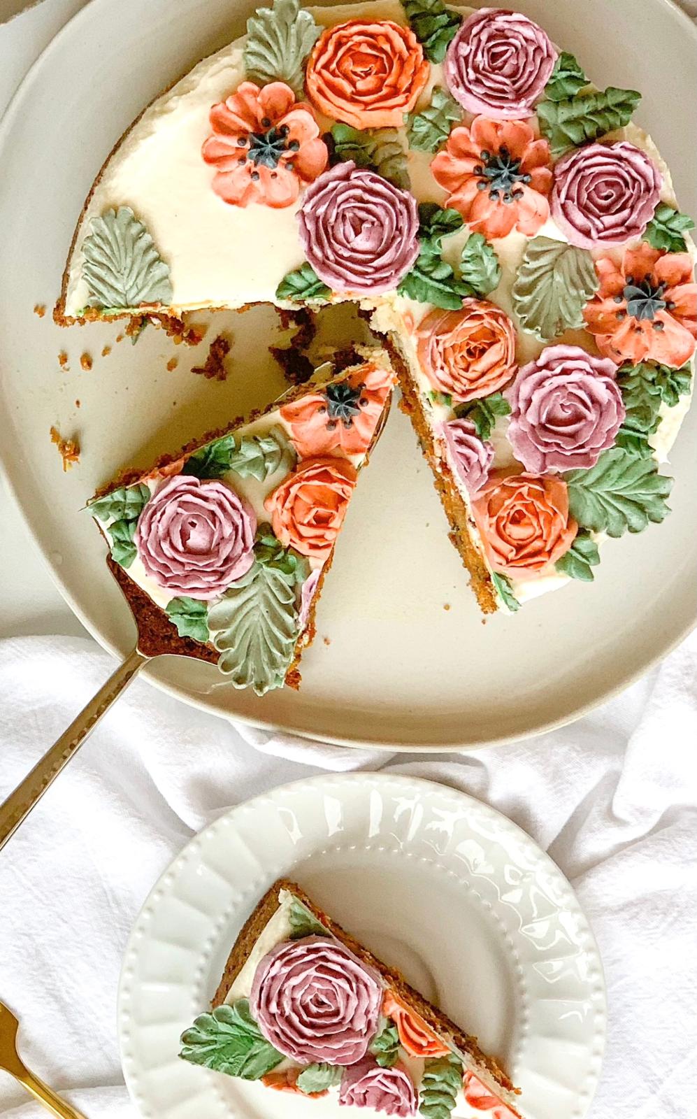 Round cake with pink and orange buttercream flowers and green leaves, with one slice cut and served on a plate.