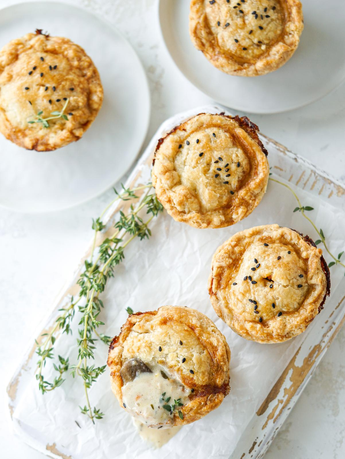 Mini savory pot pies with flaky golden crusts, black sesame seeds, and fresh thyme on parchment paper.