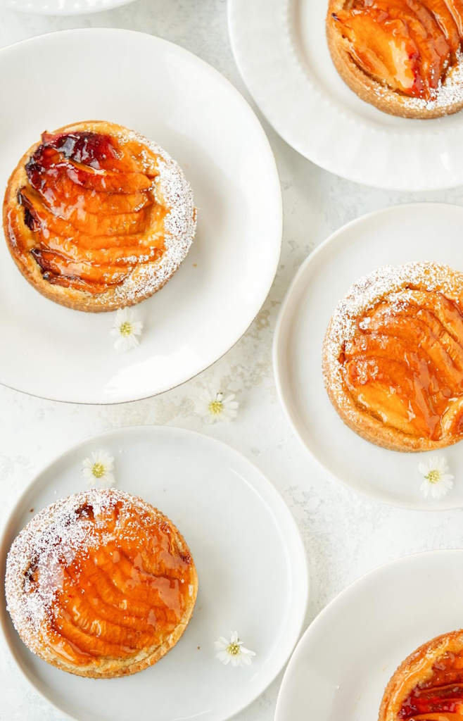 Glazed peach and almond tartlets on white plates with powdered sugar and small white flowers.