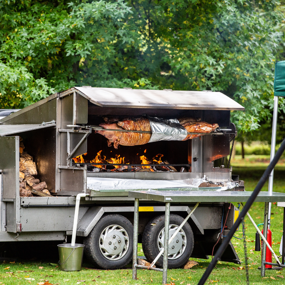 Whole pig roasting on a spit over an open flame in a large outdoor metal smoker with wood logs stored on the side.