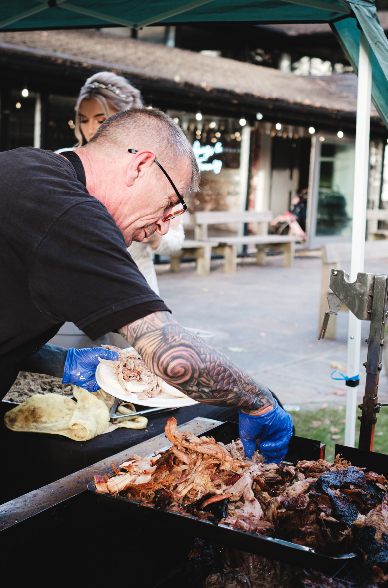 Man with tattooed arm wearing blue gloves serving pulled meat onto a plate under a canopy at an outdoor event.