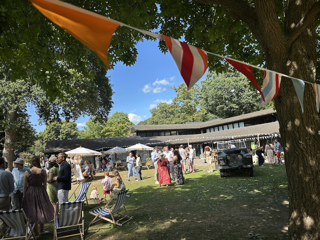 Outdoor gathering with people socializing on grass under colorful bunting and large trees beside a modern building on a sunny day.