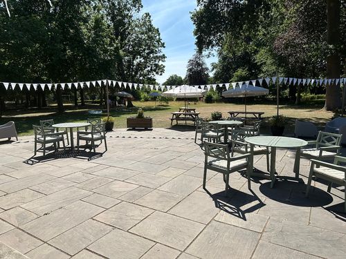 Outdoor patio with green metal tables and chairs, picnic tables with umbrellas, white triangular bunting, and trees in the background under a clear blue sky.
