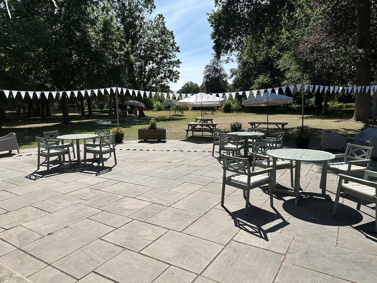 Outdoor patio with green metal tables and chairs, picnic tables with umbrellas, white triangular bunting, and trees in the background under a clear blue sky.
