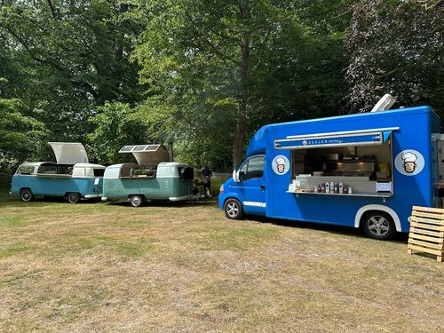Blue food truck with cartoon chef logos parked on grass next to two vintage teal camper vans with open tops, surrounded by green trees.