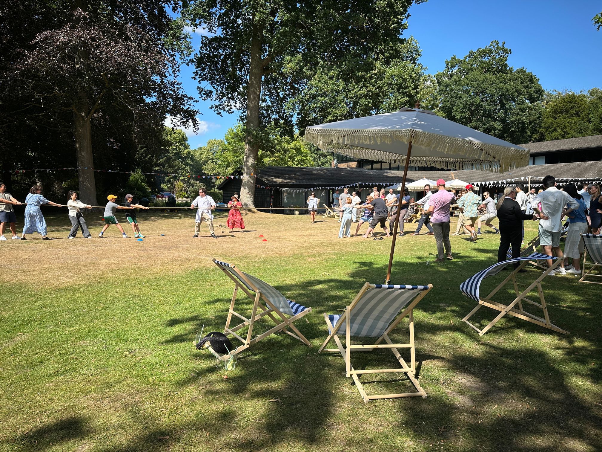 People playing tug-of-war on a sunny lawn with deck chairs and large parasols in the foreground.