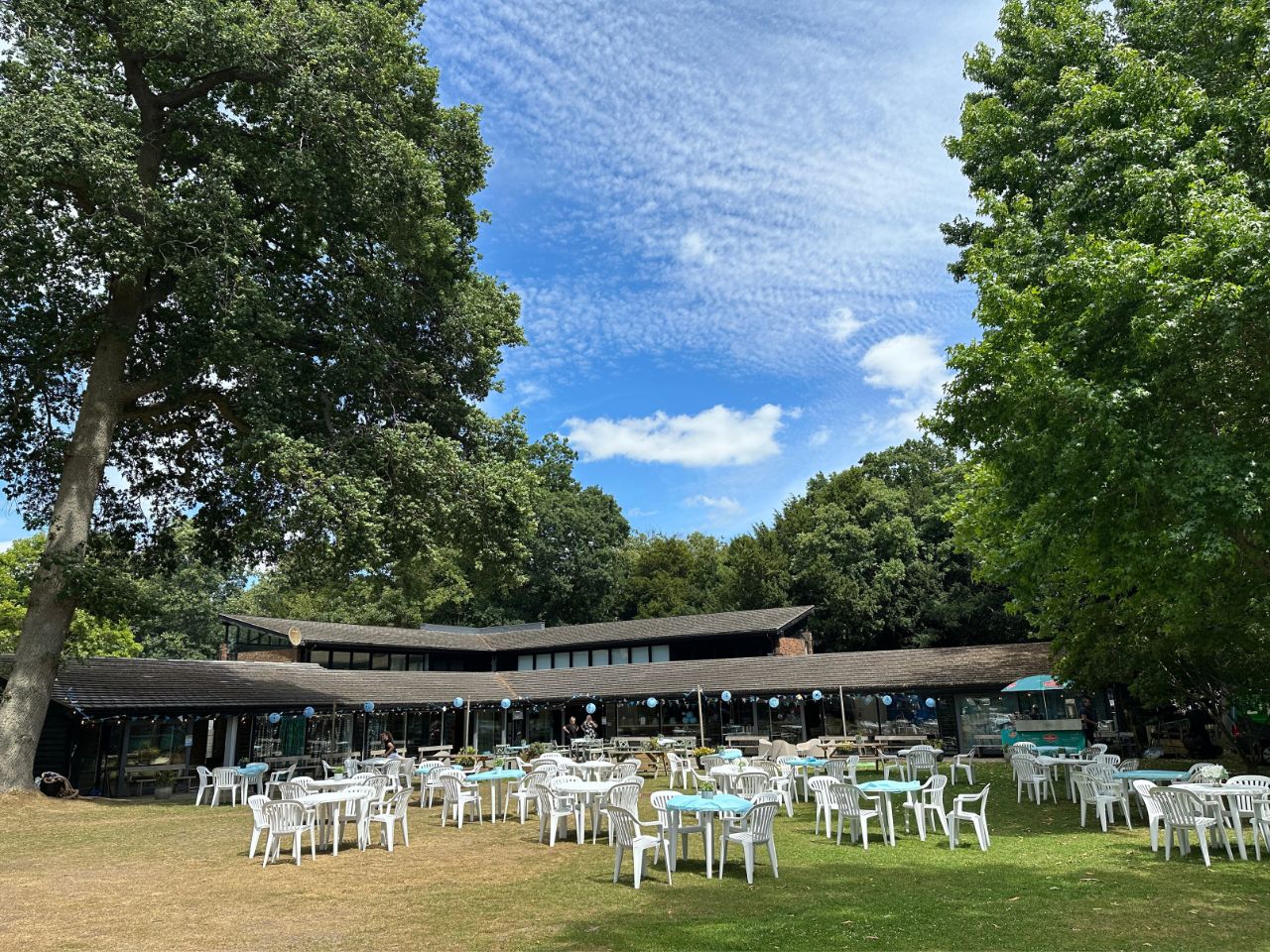 Outdoor seating area with white plastic chairs and tables with blue tablecloths on a lawn in front of a low building surrounded by large green trees under a partly cloudy blue sky.