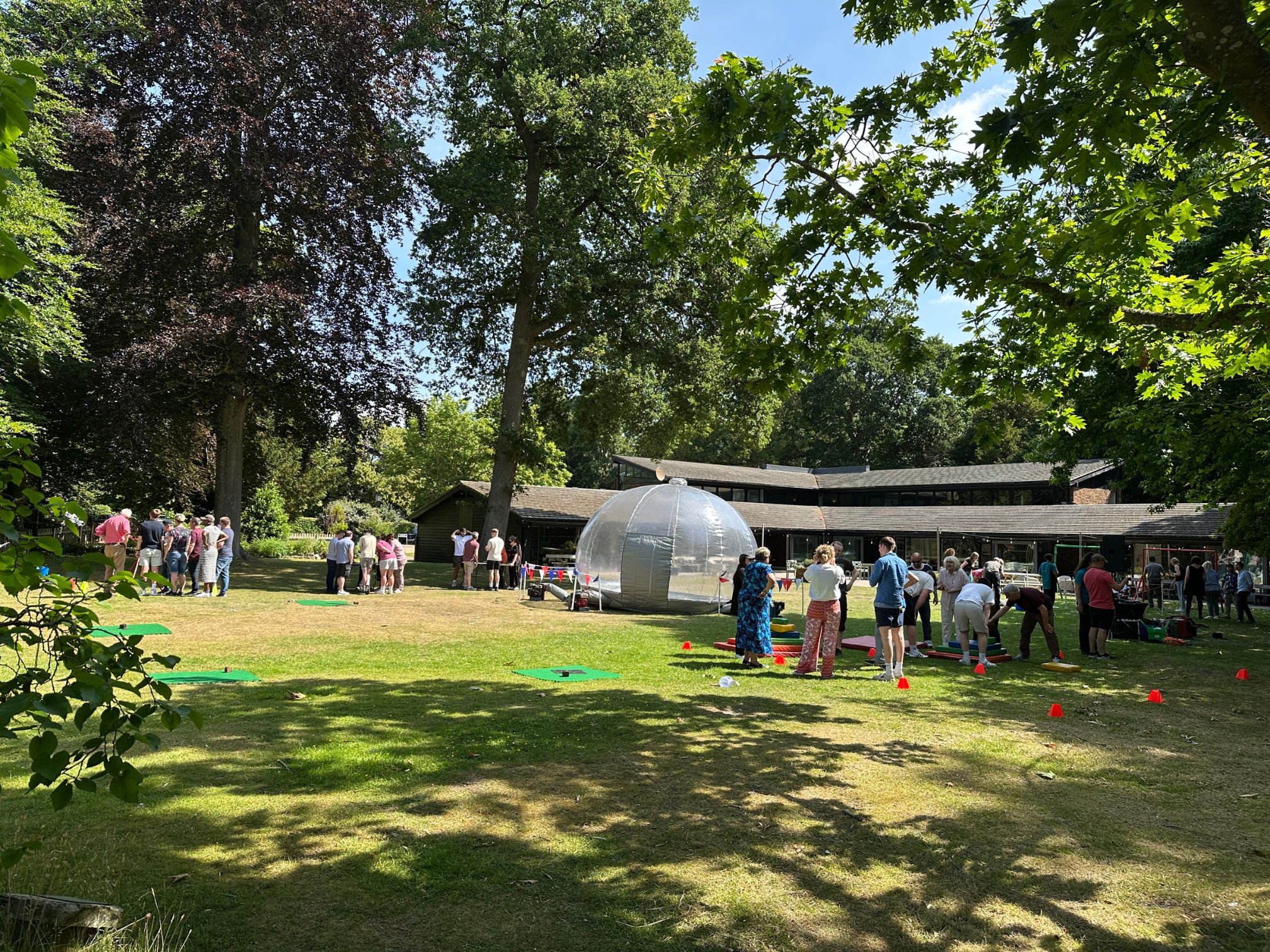 Groups of people gathered outdoors on a sunny lawn with large trees, surrounded by red cones and green mats, near a transparent inflatable dome and buildings.