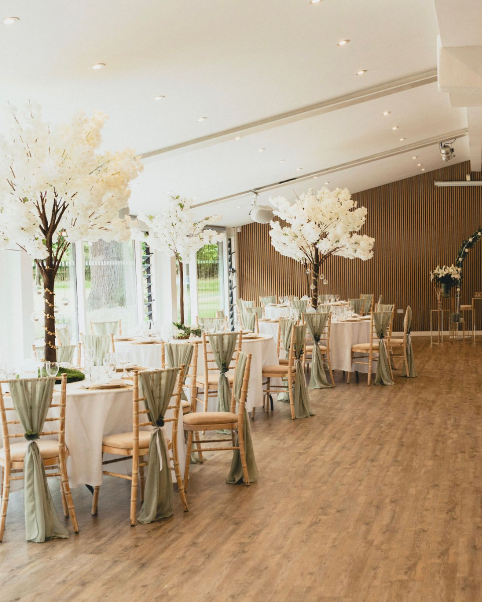 Bright event room with round tables covered in white cloths and chairs draped in soft green fabric, decorated with tall artificial white blossom trees as centerpieces.