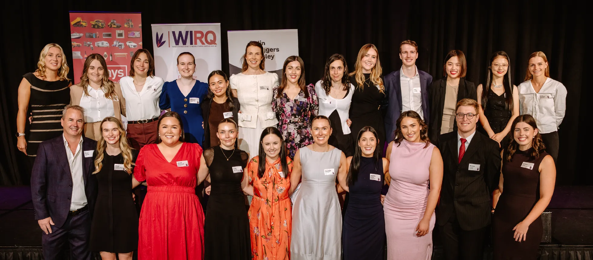 A large group of WIRQ members on stage posing for a formal committee photo in front of branded sponsor banners.