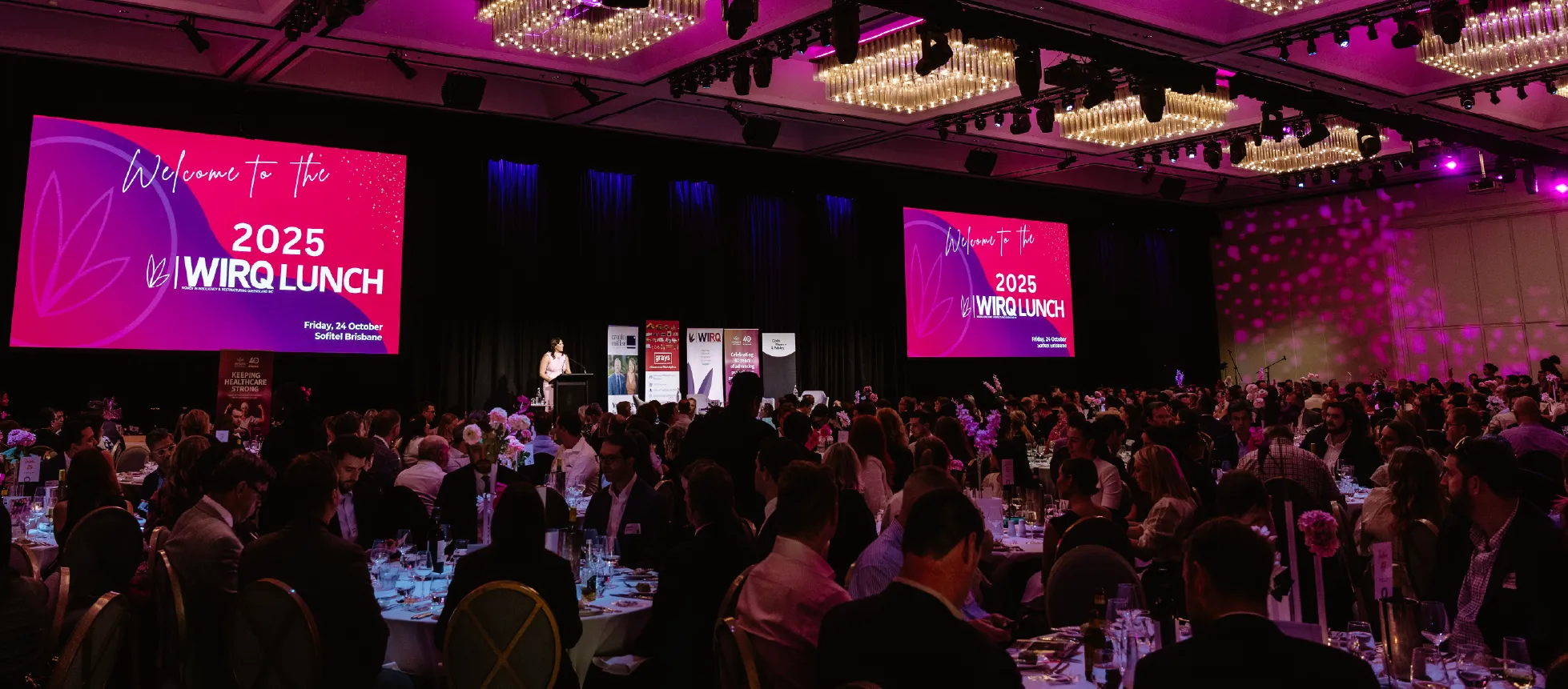 A wide shot of the WIRQ Annual Lunch with attendees seated at round tables and large event screens displaying ‘2025 WIRQ Lunch’.