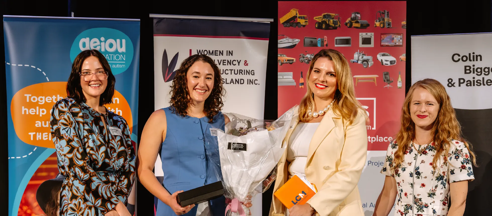 A group of WIRQ members standing together at an event, smiling in front of branded sponsor banners.
