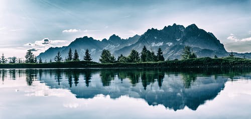 Mountain range with pine trees reflected in a calm lake under a blue sky.
