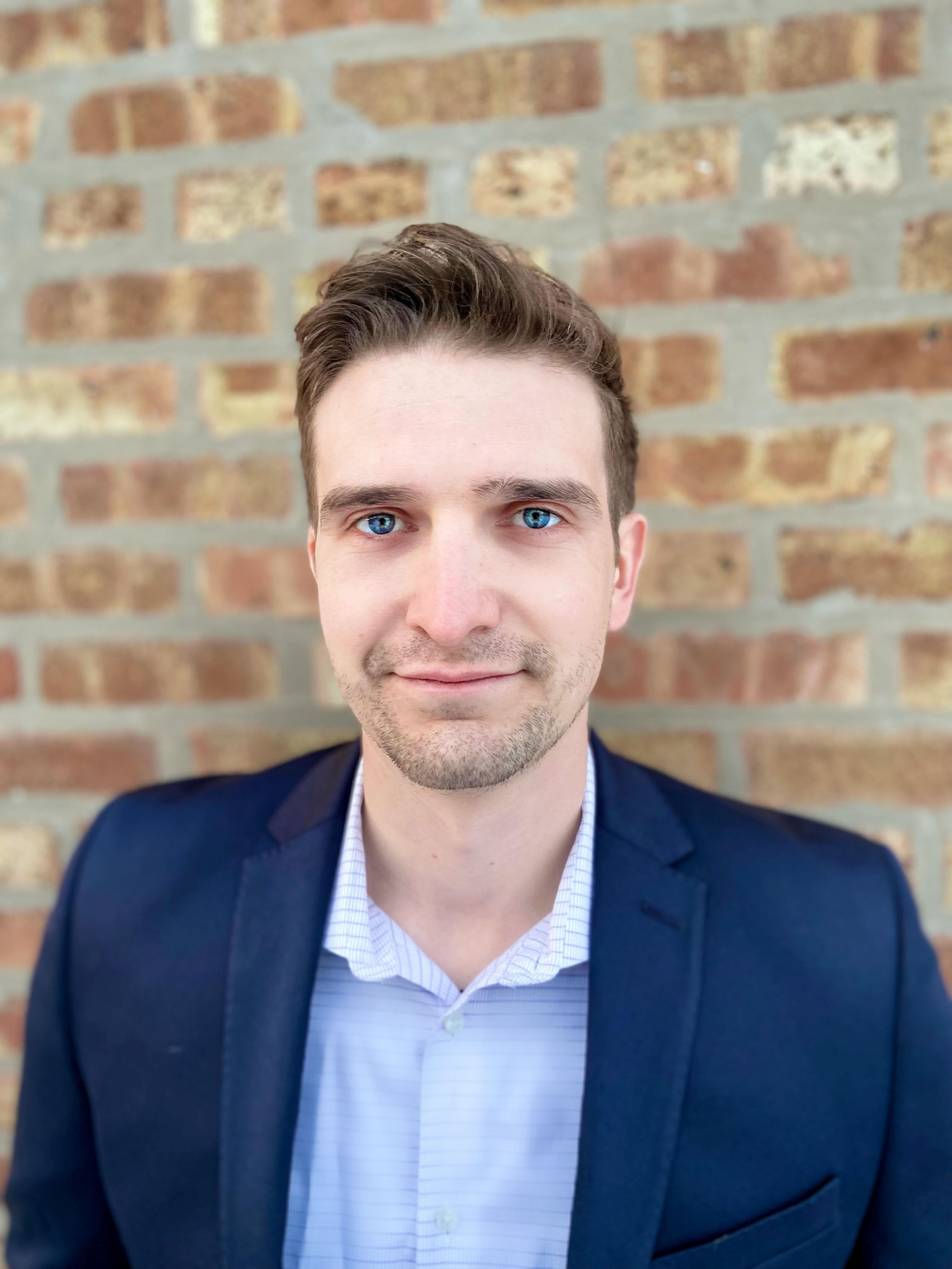 Portrait of a man with blue eyes wearing a navy blazer and light blue collared shirt standing against a brick wall.