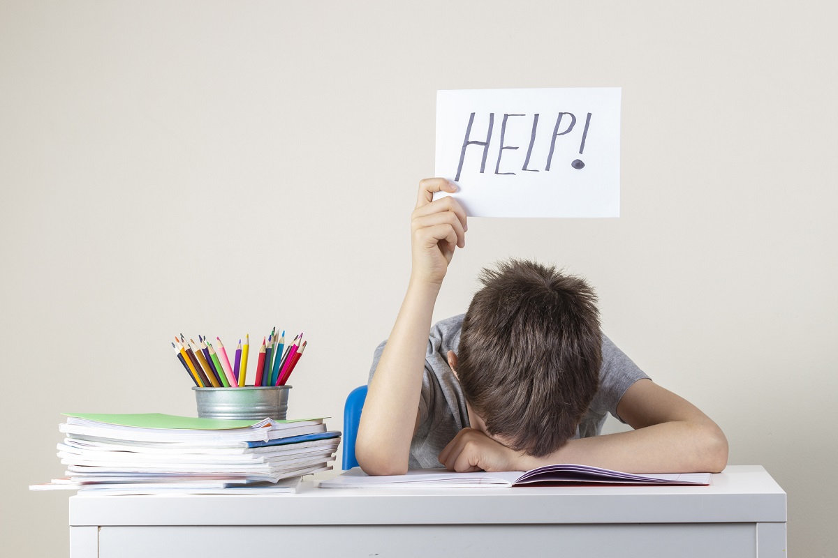 Child sitting at a desk with head down and holding a sign that says 'HELP!' next to a stack of books and colored pencils.
