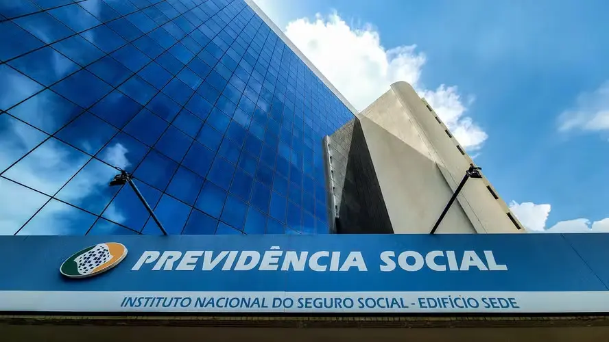 imagem principal Placa da Previdência Social em destaque na frente de um prédio espelhado com reflexo de nuvens.