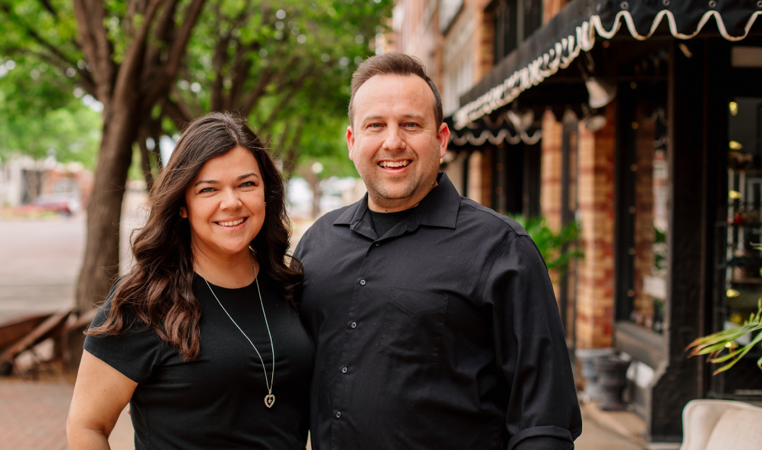 Smiling man and woman standing outdoors with trees and buildings in the blurred background.