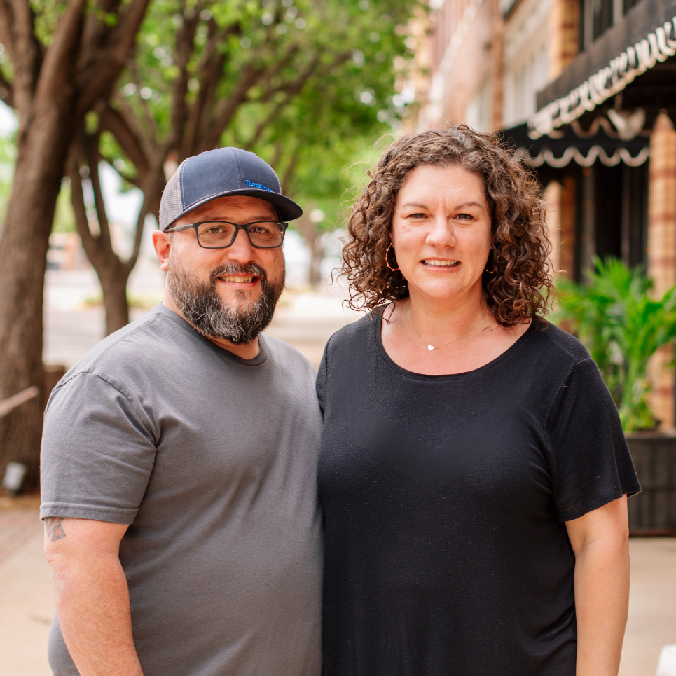 Smiling man with glasses and cap and woman with curly hair posing outdoors with trees and buildings in background.