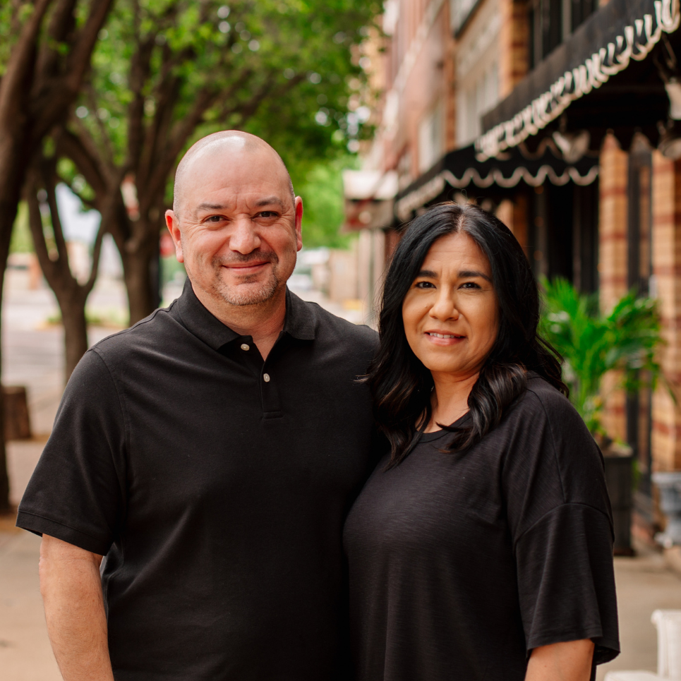 Smiling man and woman in black shirts standing side by side on a tree-lined street.