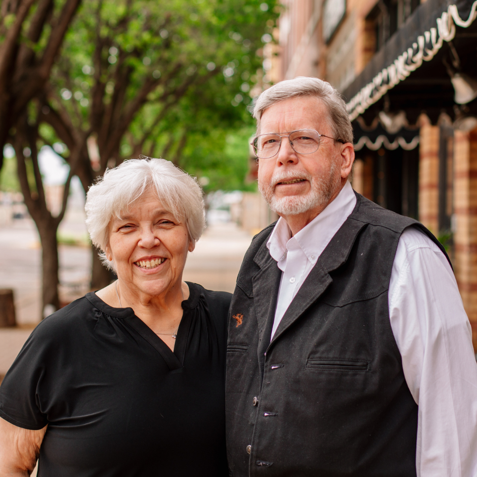 Smiling woman with white hair and man with glasses and gray beard standing outdoors.