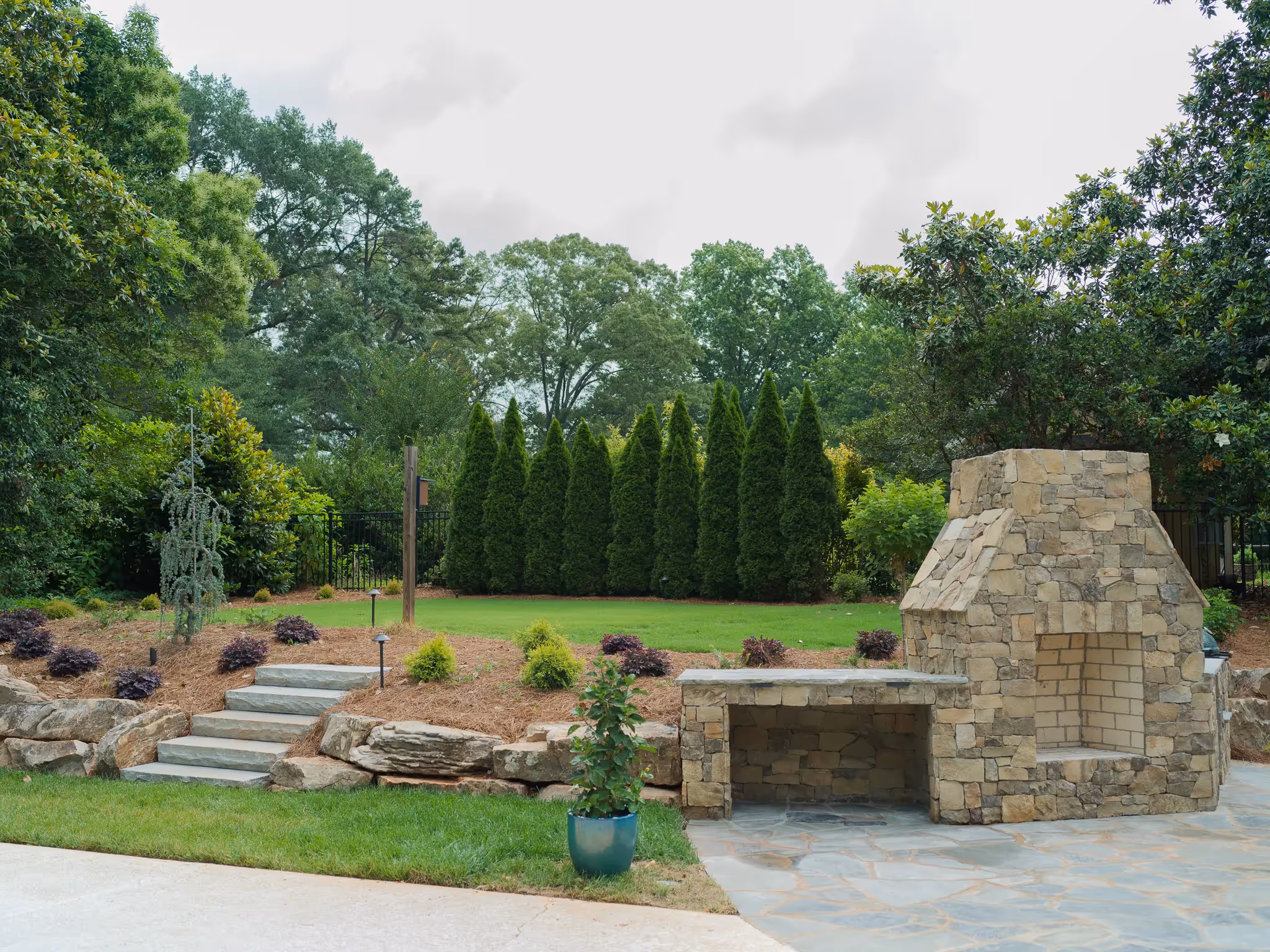Stone outdoor fireplace and built-in bench on a flagstone patio, overlooking a landscaped backyard with steps and privacy hedges