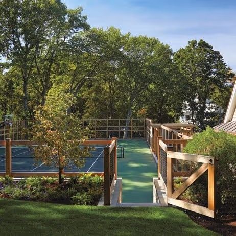 Outdoor residential pickleball court with professional blue and green surfacing, surrounded by mature trees and black chain-link fencing in a landscaped backyard setting