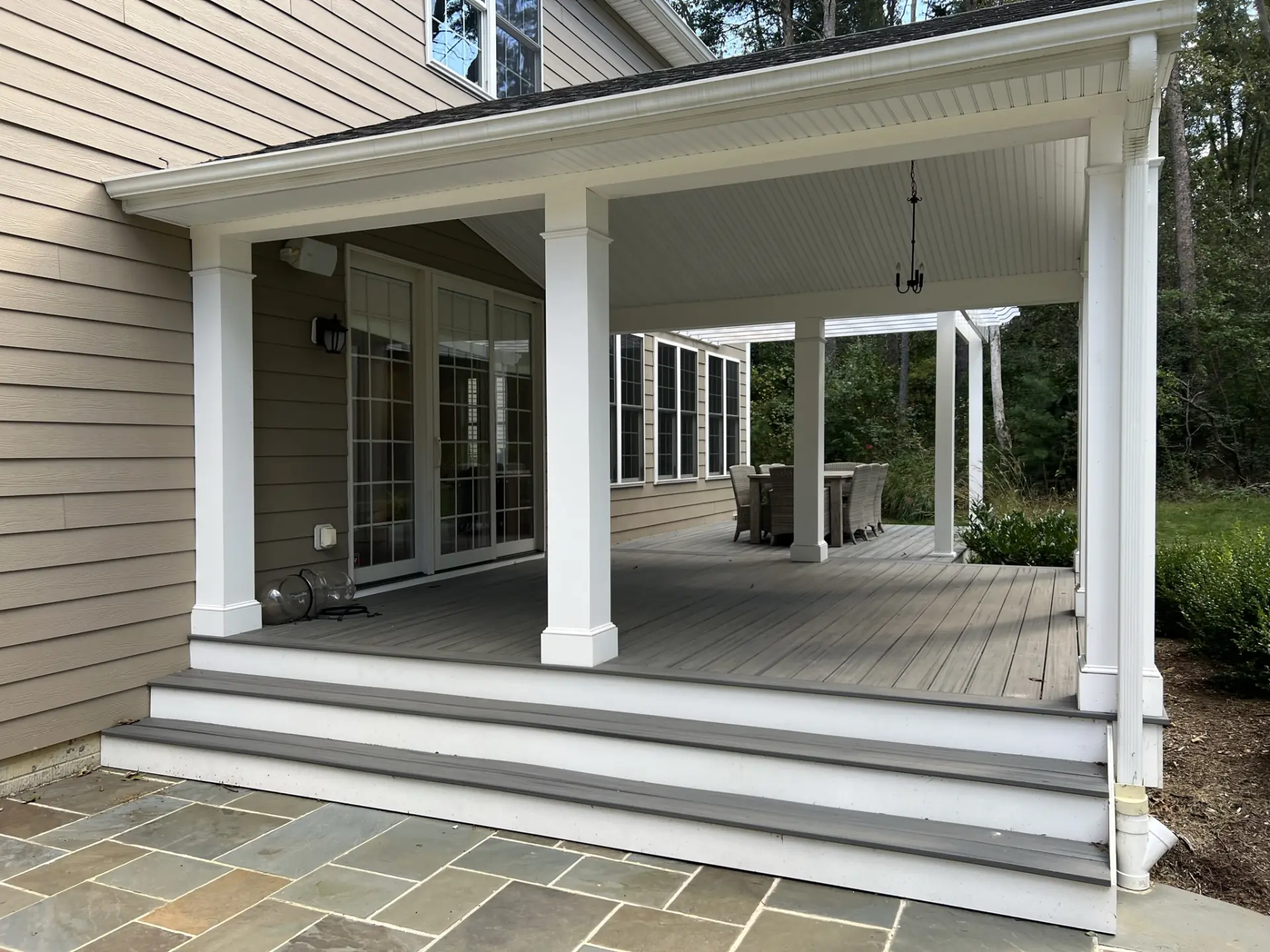 Covered composite deck attached to a beige house, featuring white columns, gray Trex or TimberTech decking, and steps leading to a stone patio, with an outdoor dining area visible in the background.